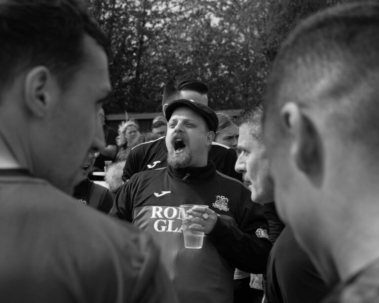 A football fan wearing a flat cap and holding a pint glass cheers, surrounded by other fans.