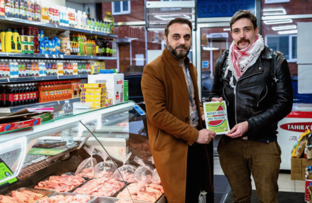 Two men, one wearing a black leather jacket and a Palestinian keffiyeh, stand next to a butcher's counter in a grocery shop.