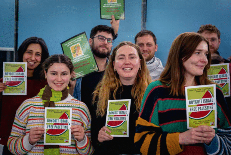 Seven activists stand in a blue hall holding green pamphlets reading 'Boycott Israel. Free Palestine'.