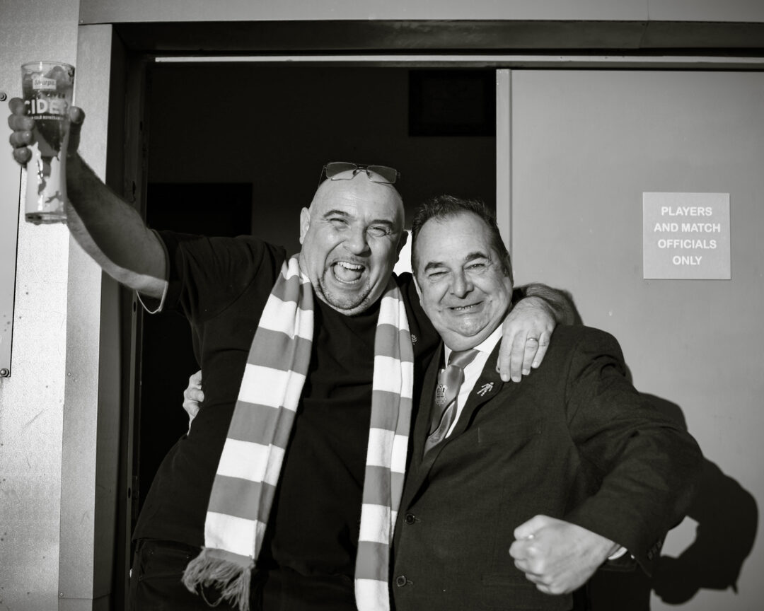 Two men, one wearing a football scarf and one wearing a suit, cheer in the doorway of a football clubhouse.