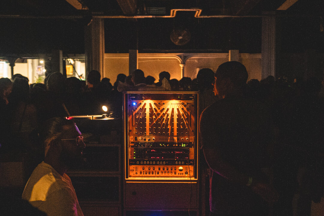 A lit up audio mixer at a sound system event, with a large crowd in between columns in the background.