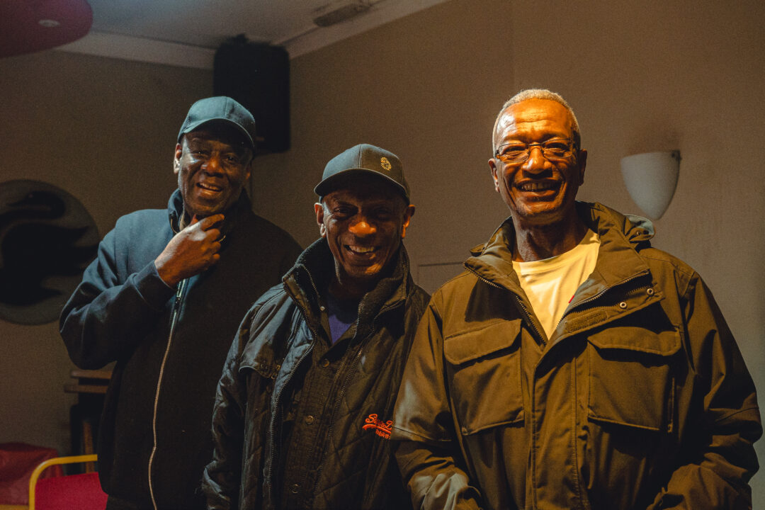 Three men in their sixties stand smiling in a room, all wearing dark coats.