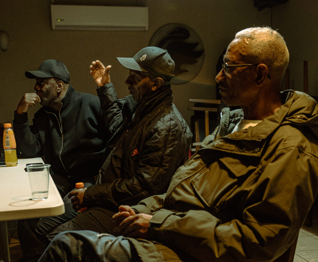Three men in their sixties sit at a table. Two wear black caps and one wears a duffle coat.