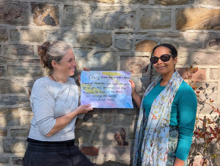 Two women hold a sign between them against a brick wall