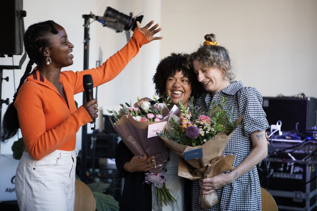 Two women, both holding bunches of flowers, laugh while another woman with a microphone gestures next to them. 