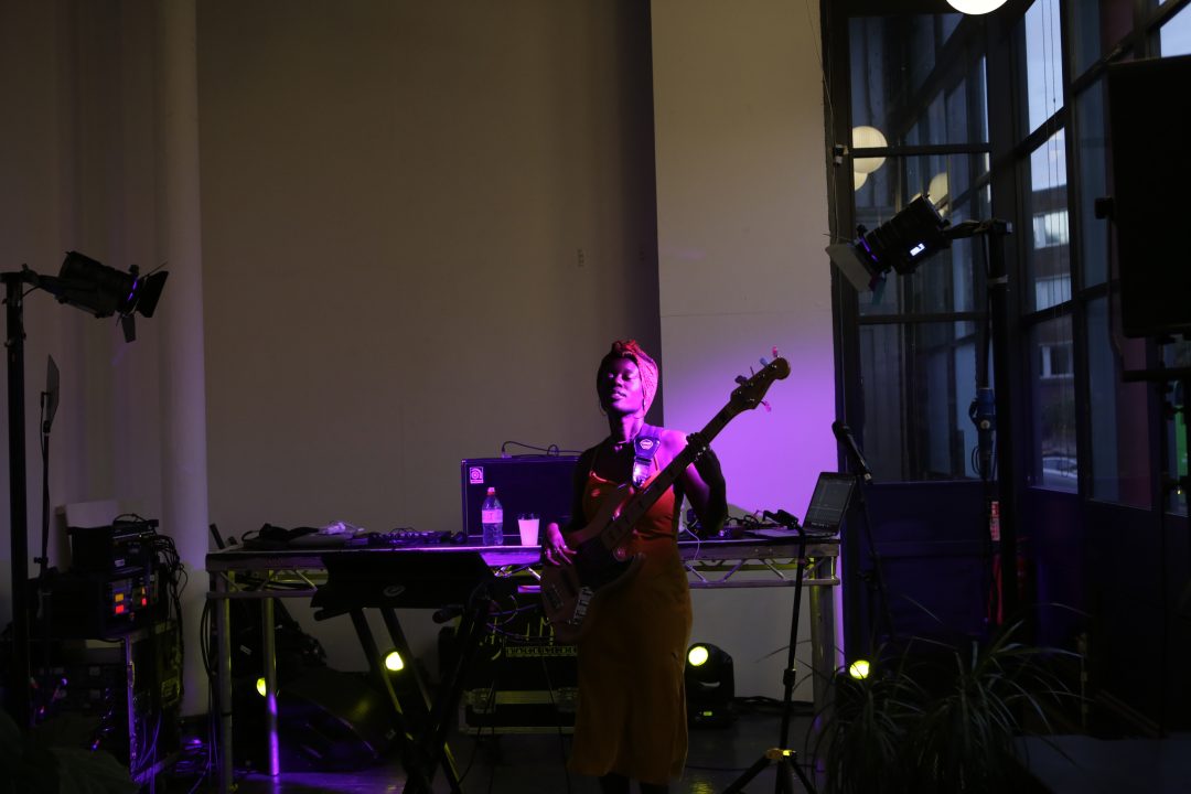 A woman plays the bass guitar in a darkened room, illuminated by purple light. 