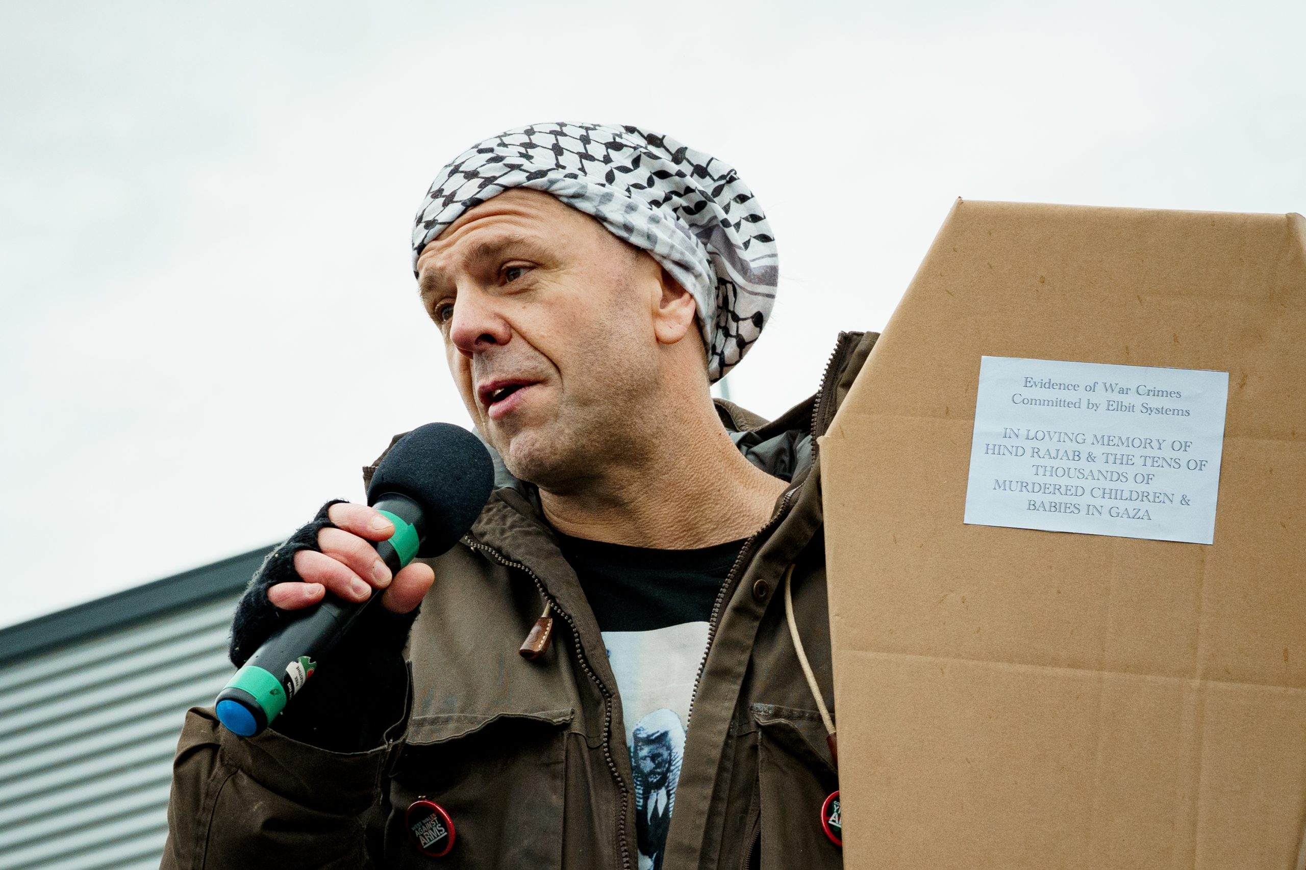 A man wearing a green jacket and holding a cardboard coffin speaks into a microphone