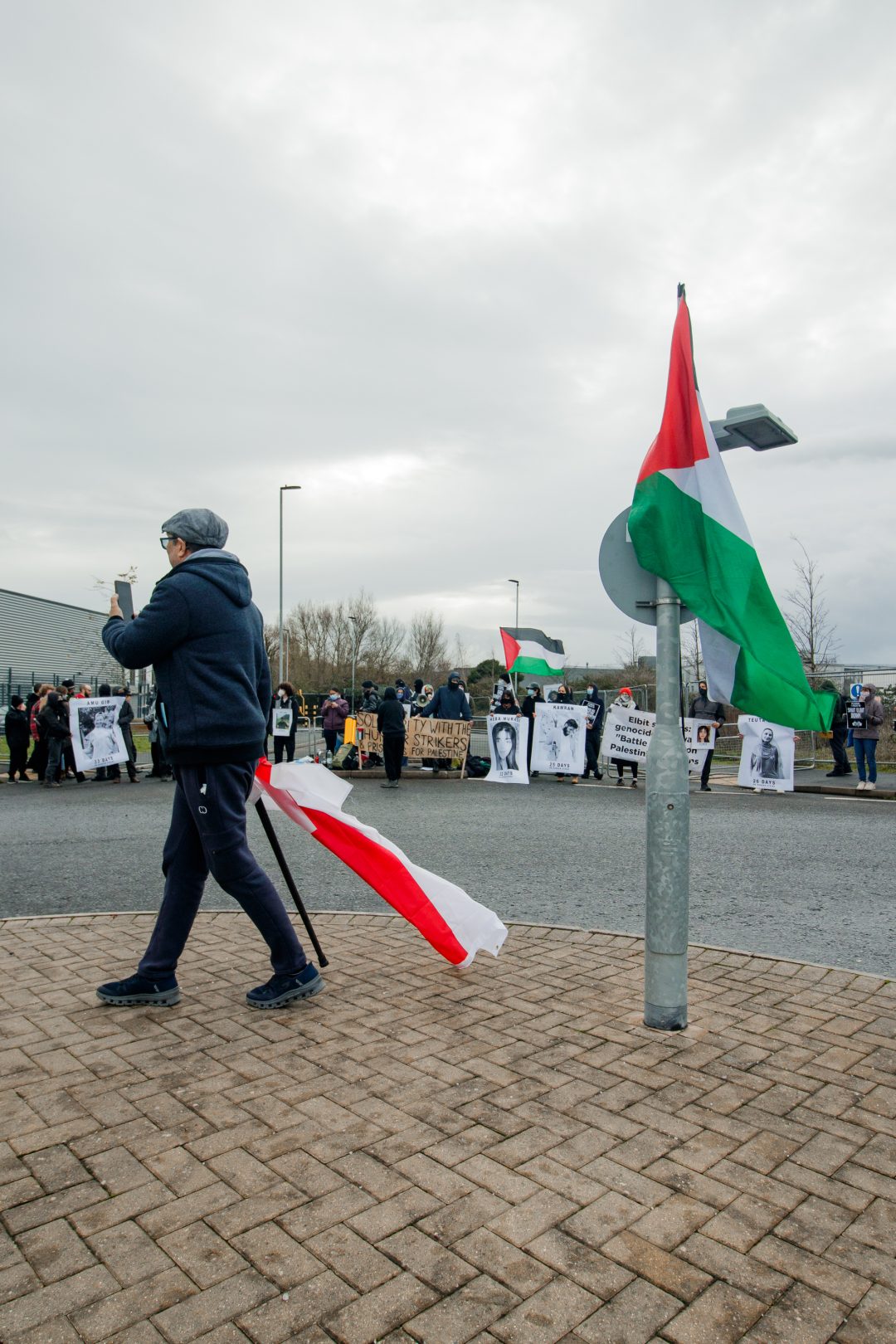 A man with a walking stick and and English flag films a group of protesters on his phone. 
