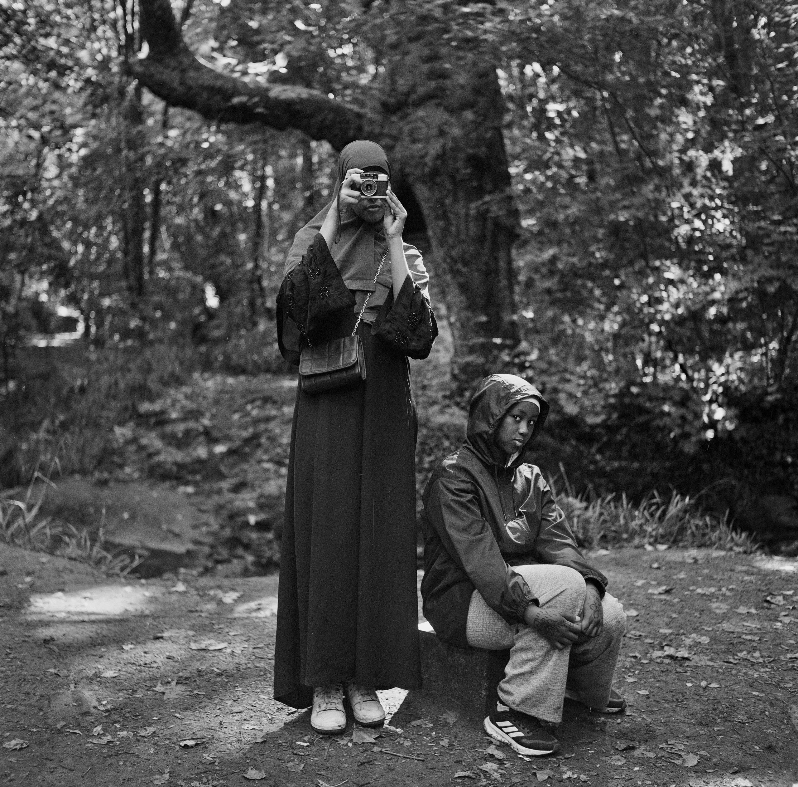 A girl stands with a camera next to a girl sitting on a block in a park.
