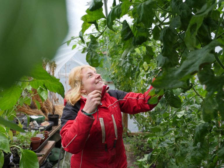 A smiling woman in a red jacket works in a greenhouse surrounded by plants.