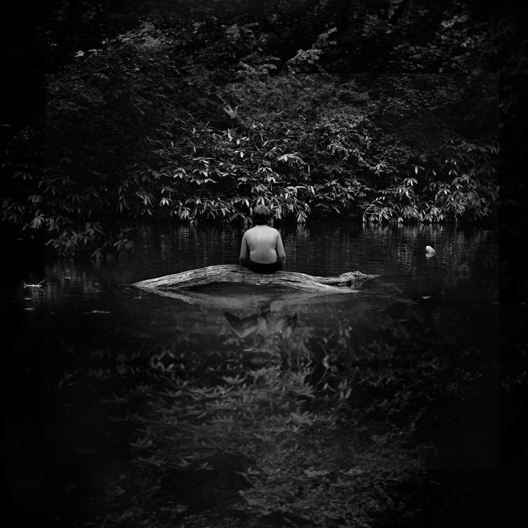 A boy sits alone on a log in a river. 