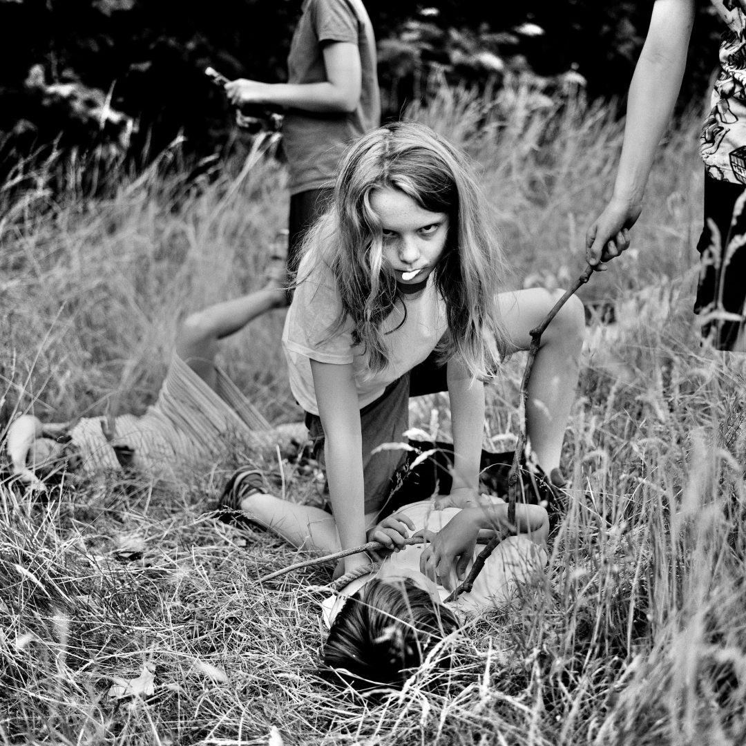 A girl kneels over a boy, among a group of other children playing a park. 