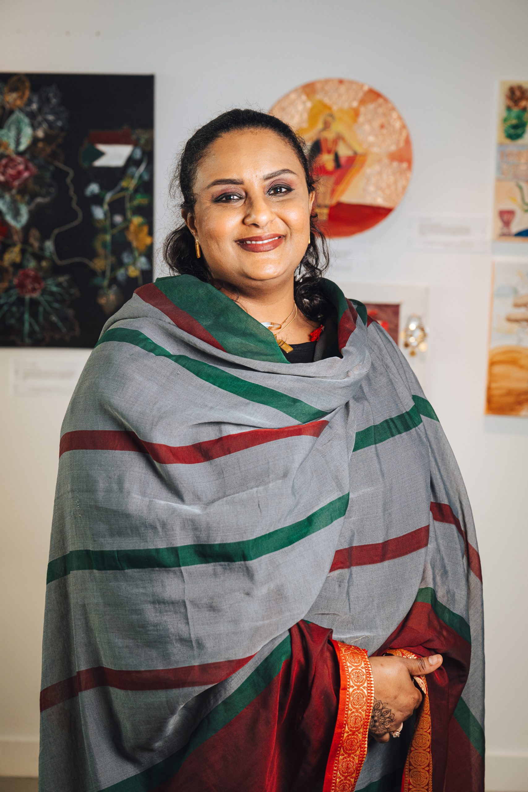 A smiling woman standing in front of artworks on a wall, wearing a grey, green and red shawl