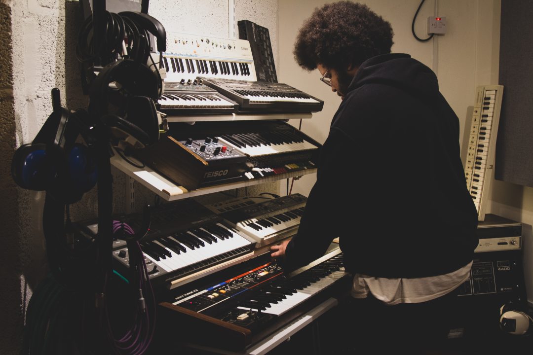 A man wearing a black hooded jumper stands in front of a rack of keyboards in a music studio