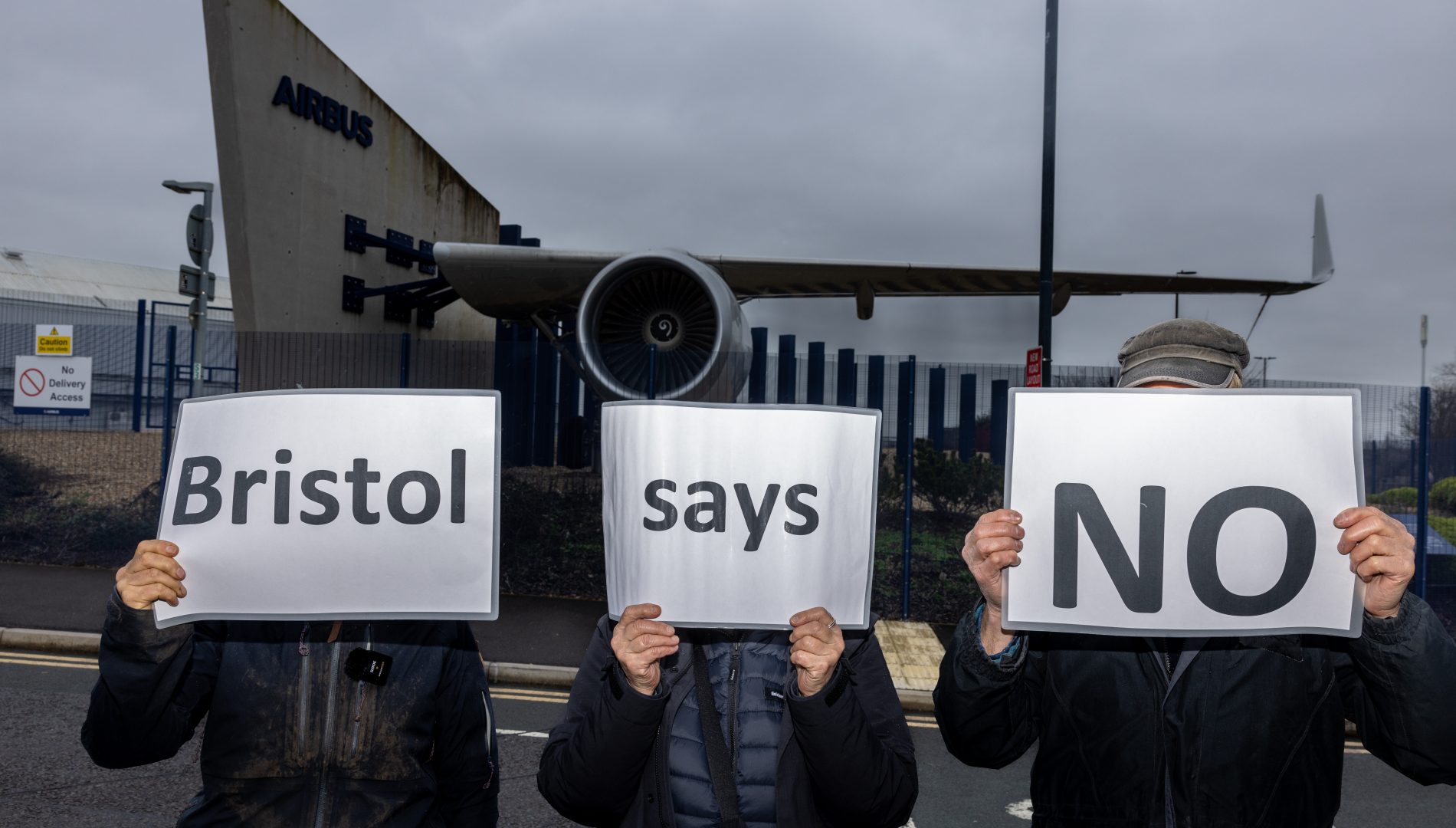 Three people standing in front of a jet wing hold white signs in front of their faces