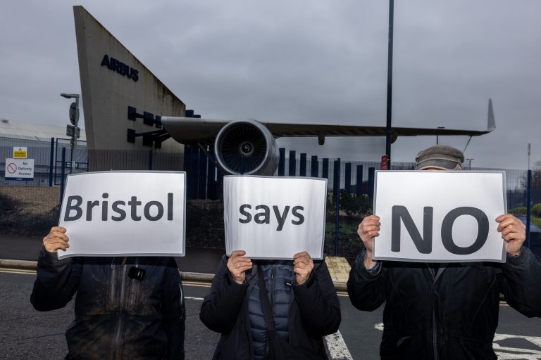 Three people standing in front of a jet wing hold white signs in front of their faces
