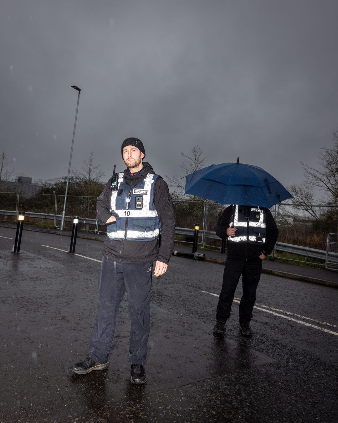 Two security guards wearing high-viz jackets stand in the road on a rainy day. 