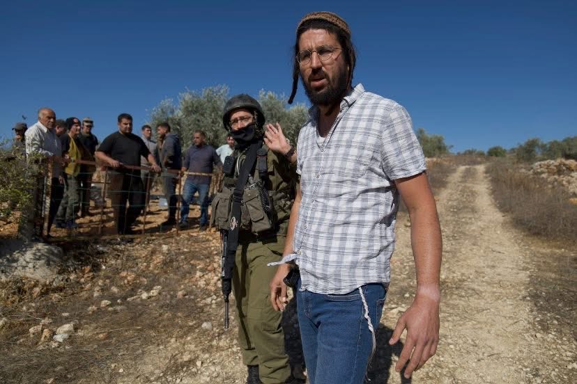 A white in a light shirt stands next to a solider before a fence where several other men stand. 