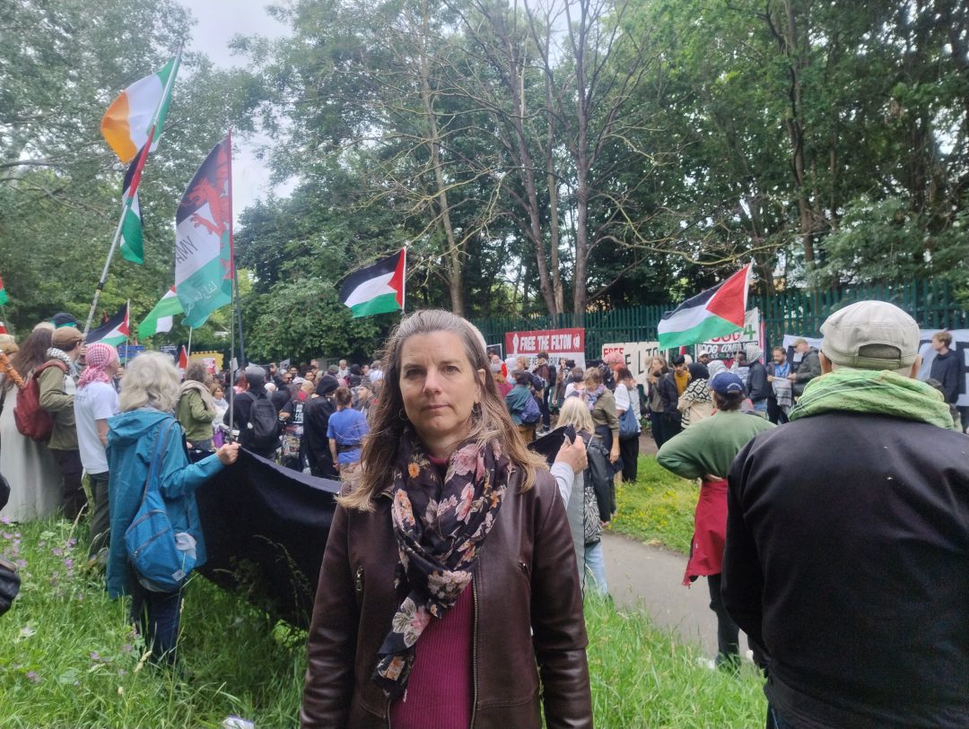 A woman wearing a brown leather jacket and scarf stands in front of a crowd of people holding banners and waving flags. 