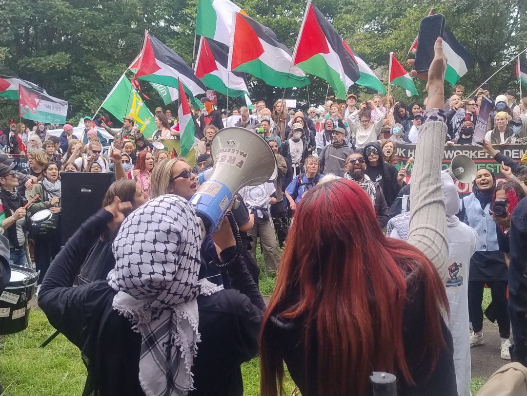 Large group of protesters chanting and waving Palestine flags in front of a row of trees.