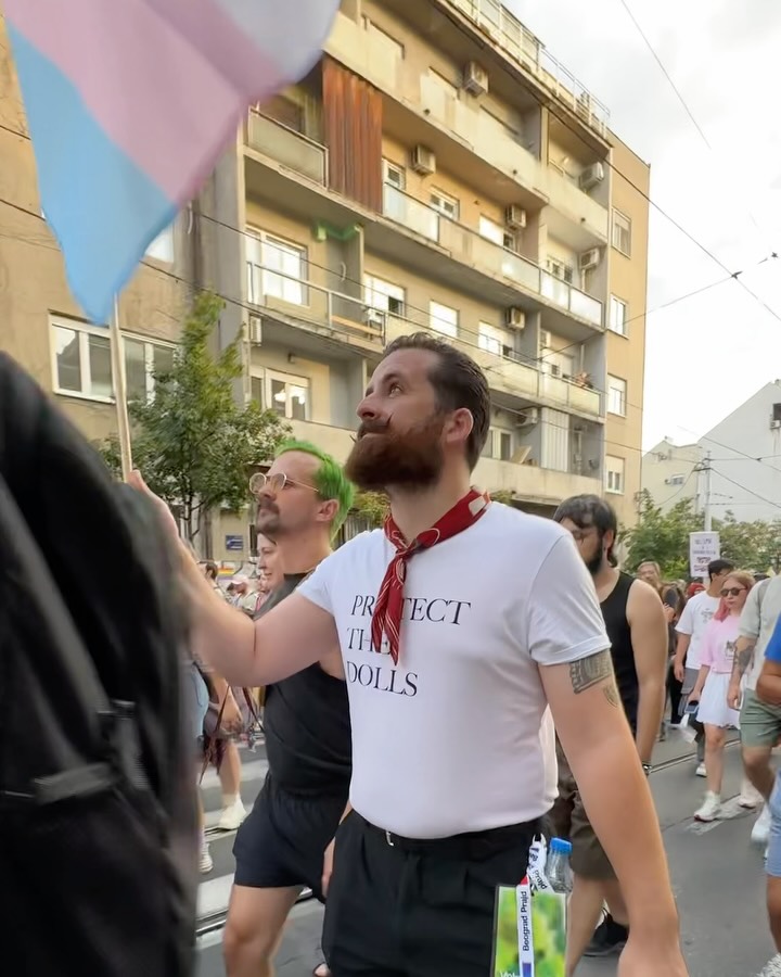A man wearing a white t shirt and a red bandana carrying a pride flag as marchers walk down a city street