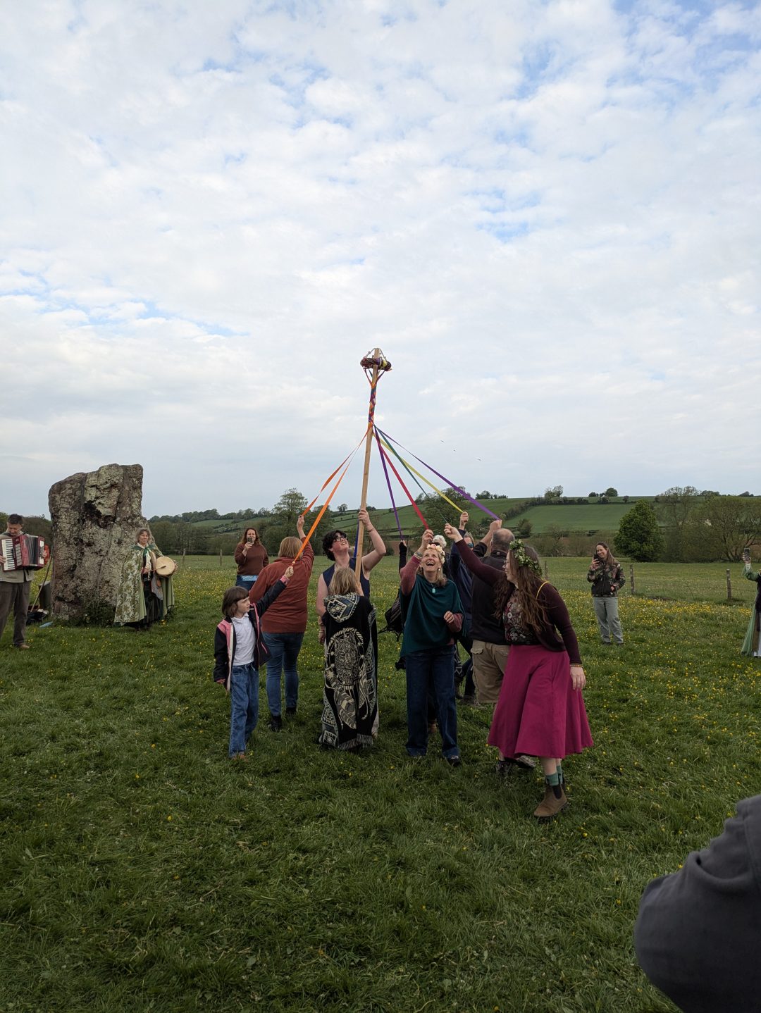 A group of people dance around a pole in a field at the edge of a stone circle