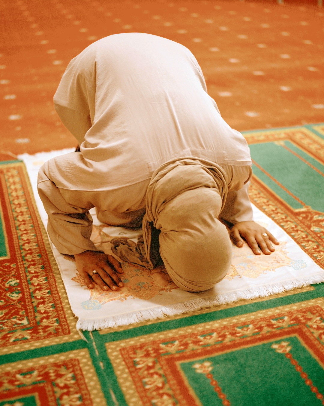 A young woman wearing beige clothes prays on an orange carpet