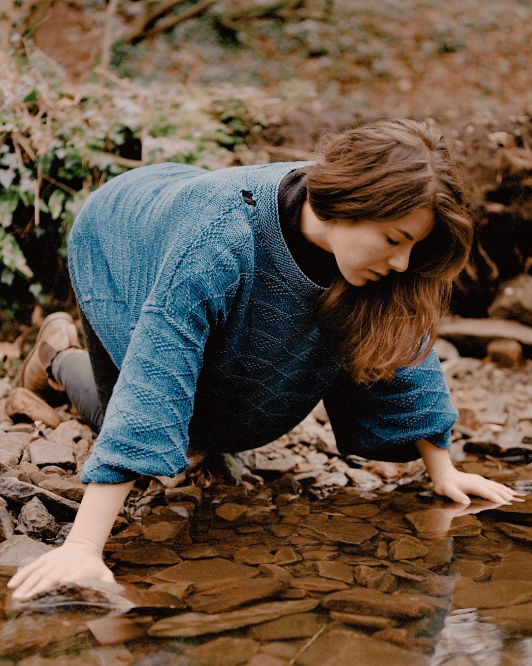 A woman kneels close to the edge of a body of water with her hands on some rocks