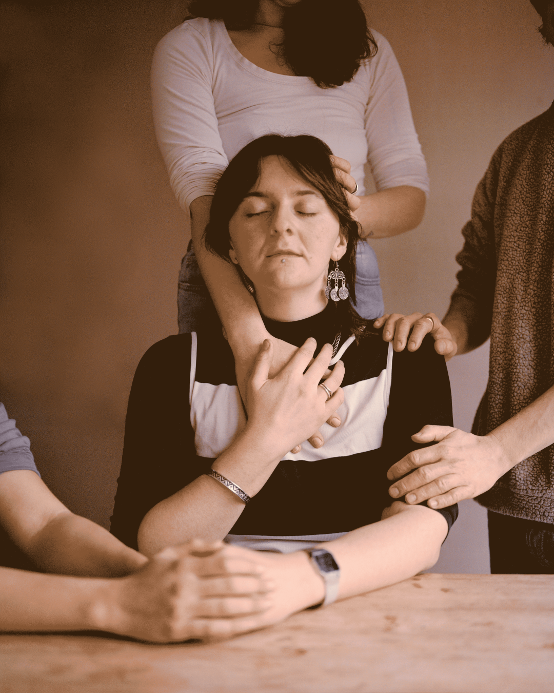 A young woman with her eyes closed sits at a table surrounded by friends gently placing their hands on her