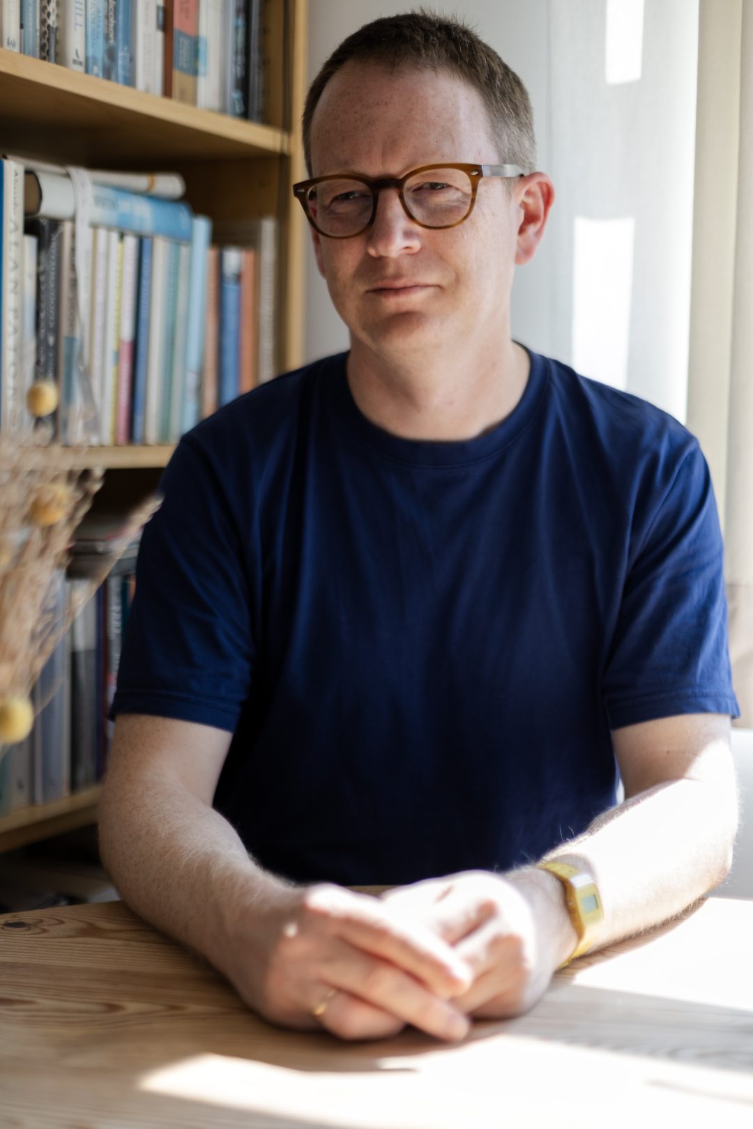 A man in a blue t-shirt and glasses sits at a wooden desk next to a bookshelf
