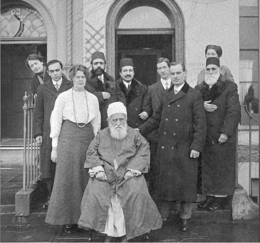 Black and white photo of a seated man in a robe and white turban with a group of men and women in smart dress standing behind