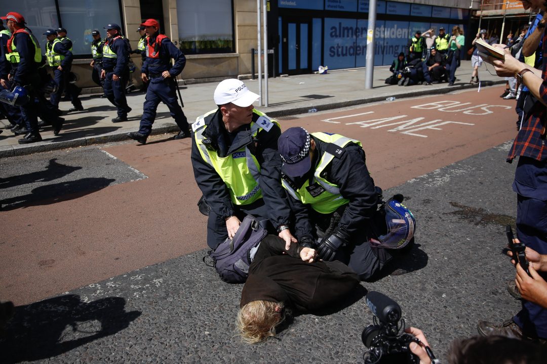Two police officers wearing caps and fluorescent jackets handcuff a protester who is face down on the road