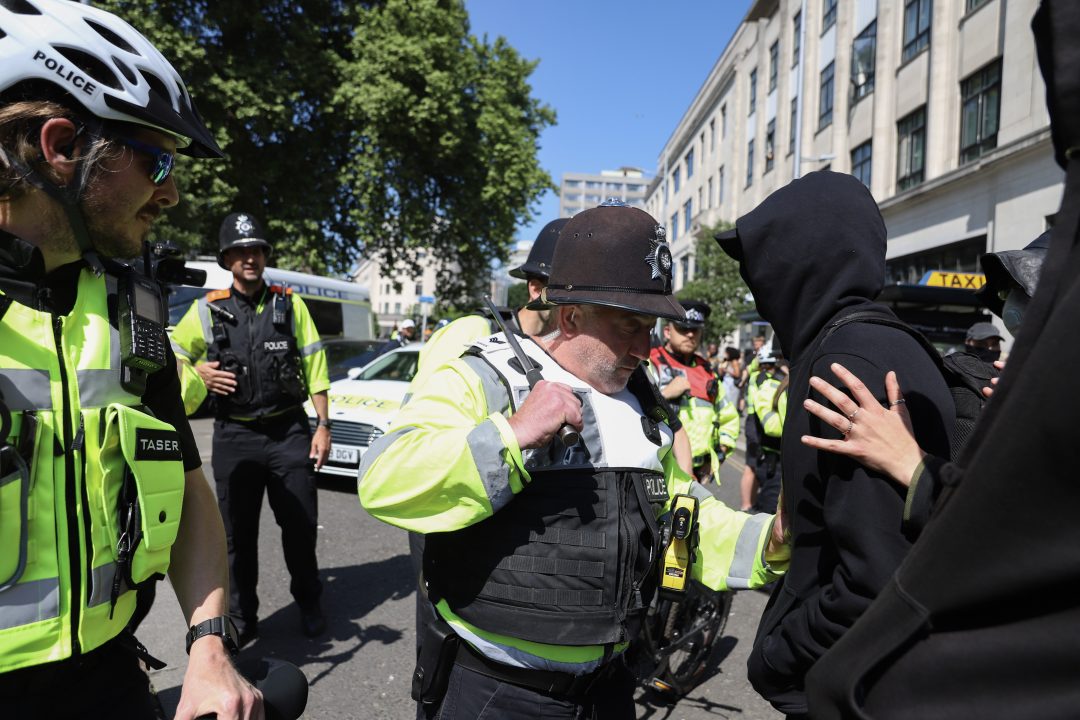 A police officer with his baton drawn stands in front of a masked protester wearing all black