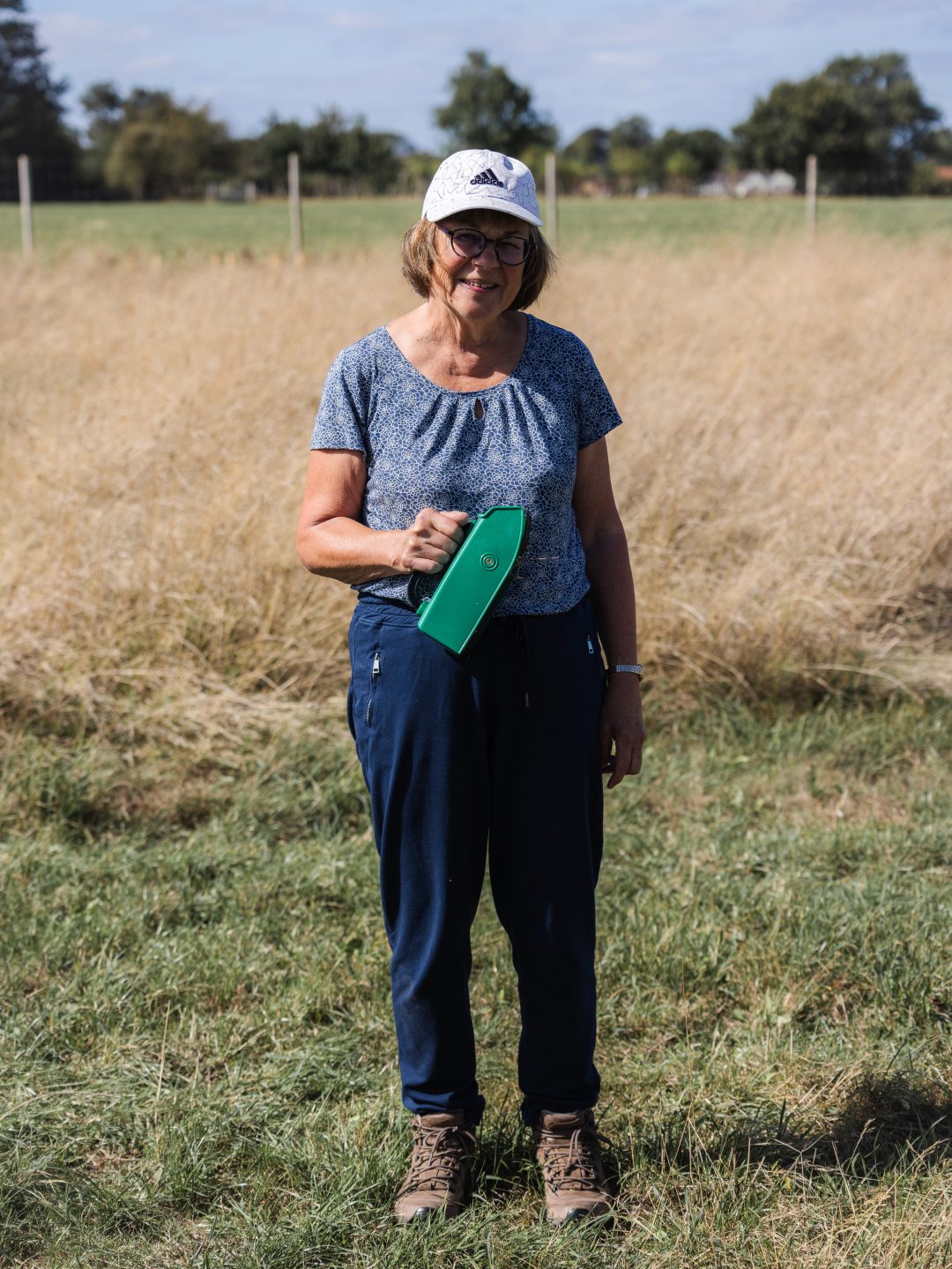 A woman in a white baseball cap and blue trousers stands holding a small green container in a field