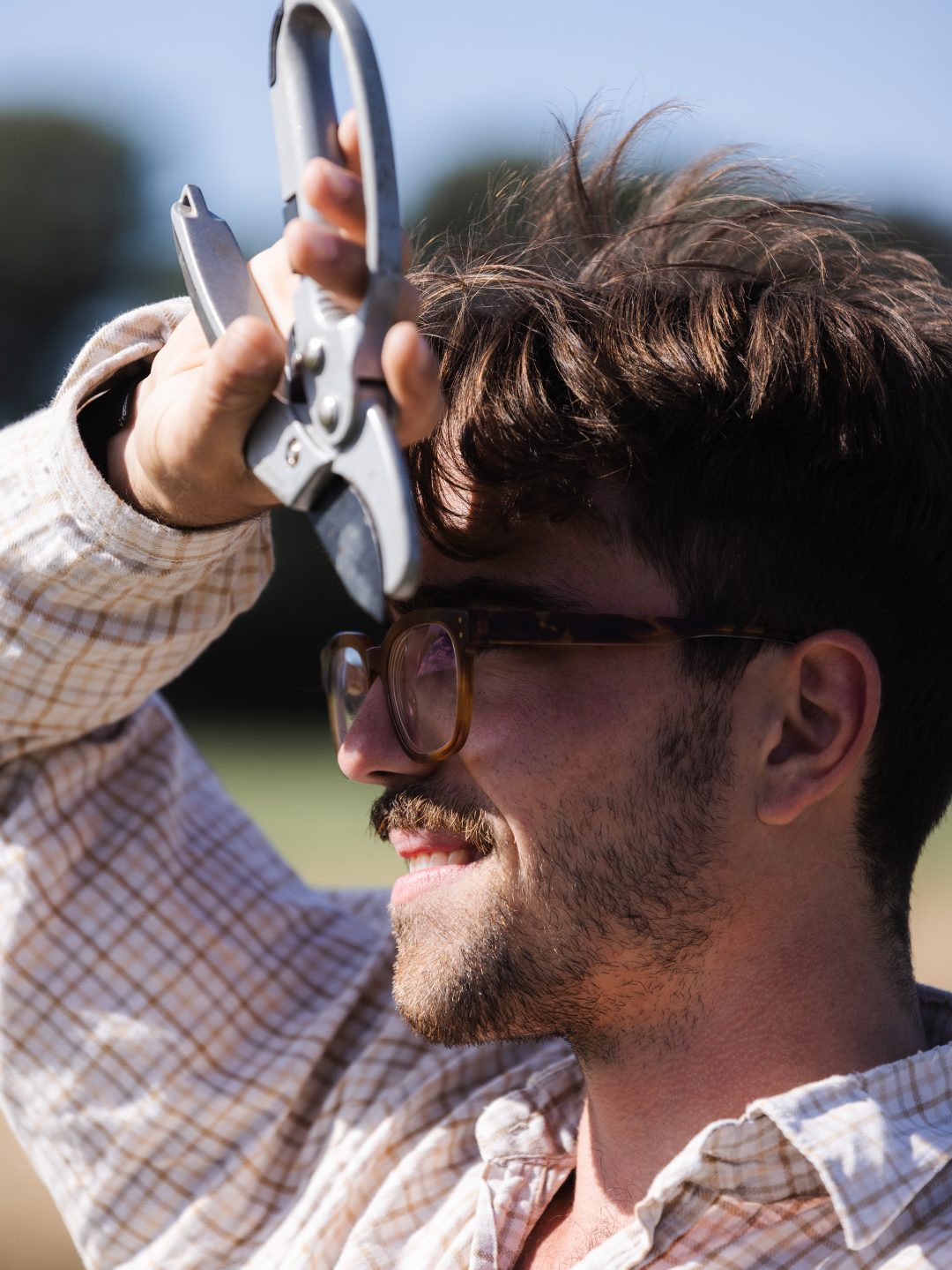 A man in a white striped shirt and glasses holds a pair of pruners against his head to shield his eyes from the sun