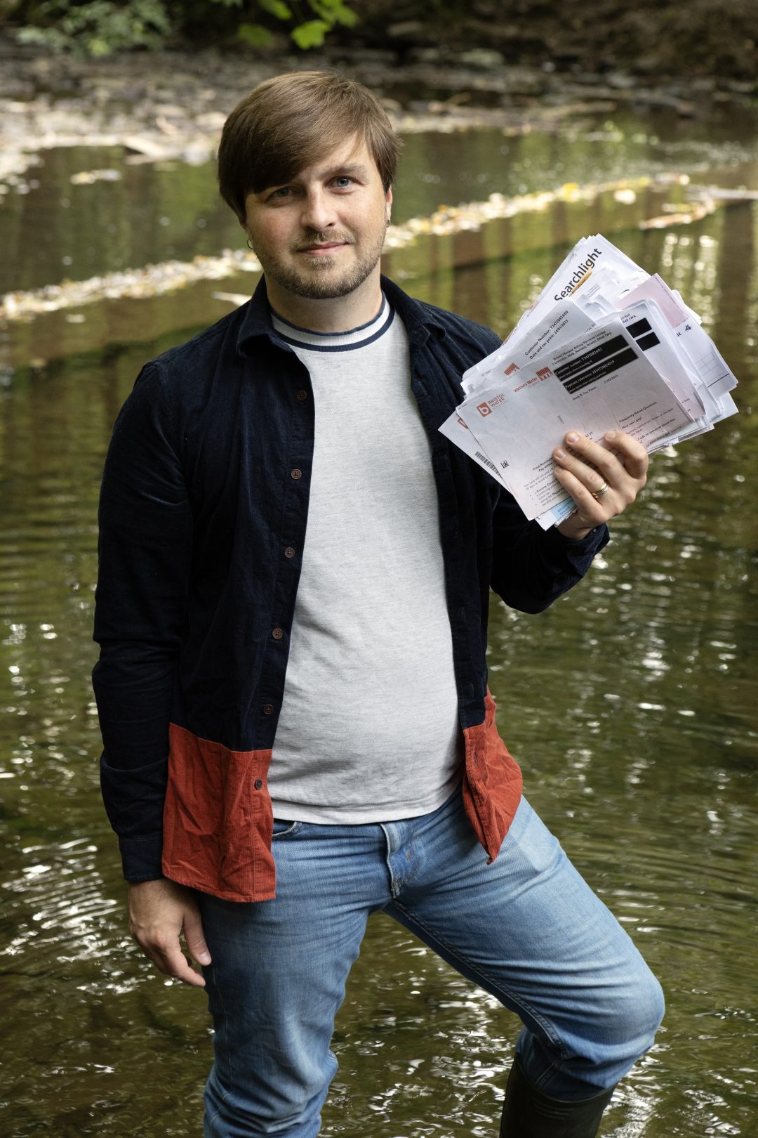 A man in a blue shirt and jeans stands on the banks of a river while holding a large pile of letters 