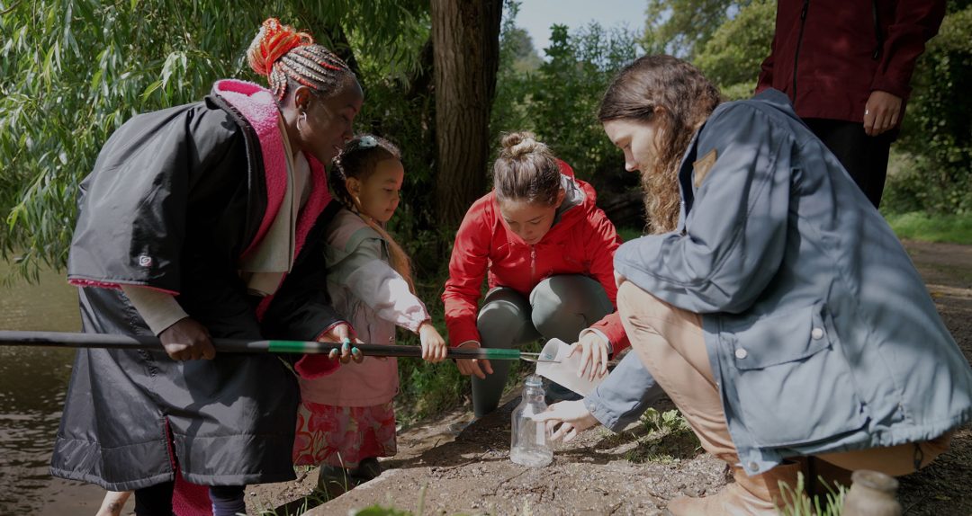 A group of three women and a young girl pouring river water into a jar on a muddy riverbank 