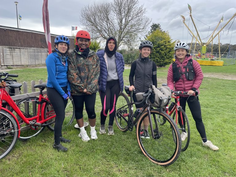 Five women wearing bike helmets and holding bikes in a small park