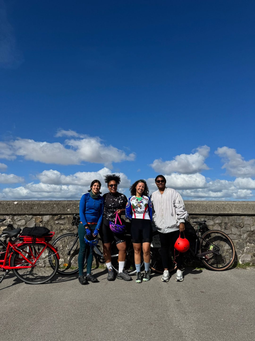 Four women leaning against a concrete wall with their bicycles, with a wide blue sky above